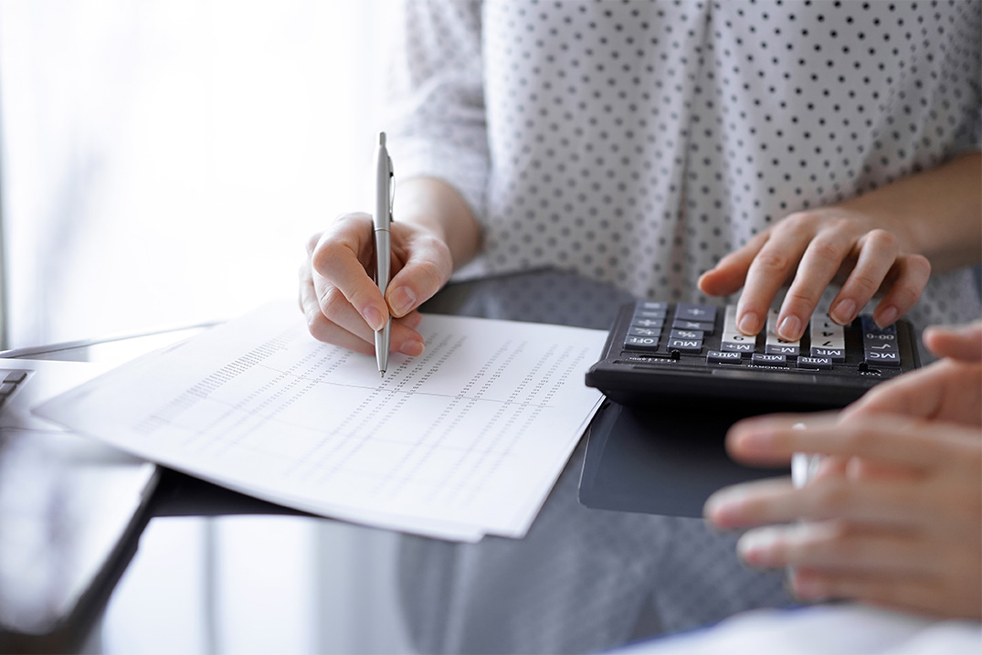 Nahaufname von Händen. Rechte Hand zeigt mit dem Kugelschreiber auf ein Dokument. Linke Hand tippt auf einem Taschenrechner.